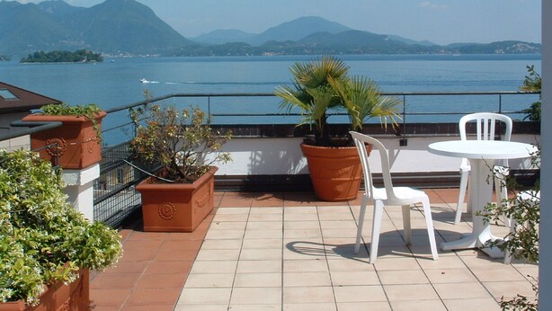 Terraço com plantas em vasos, uma cadeira branca e uma mesa com vista para um lago e montanhas ao fundo.