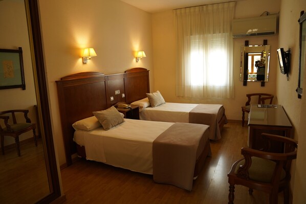A hotel room with two single beds, a wooden nightstand, a desk with a chair, a mirror, and a window with curtains.