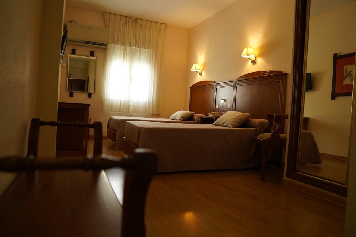 A hotel room with two beds, a desk, a chair, a mirror, and a window with curtains.