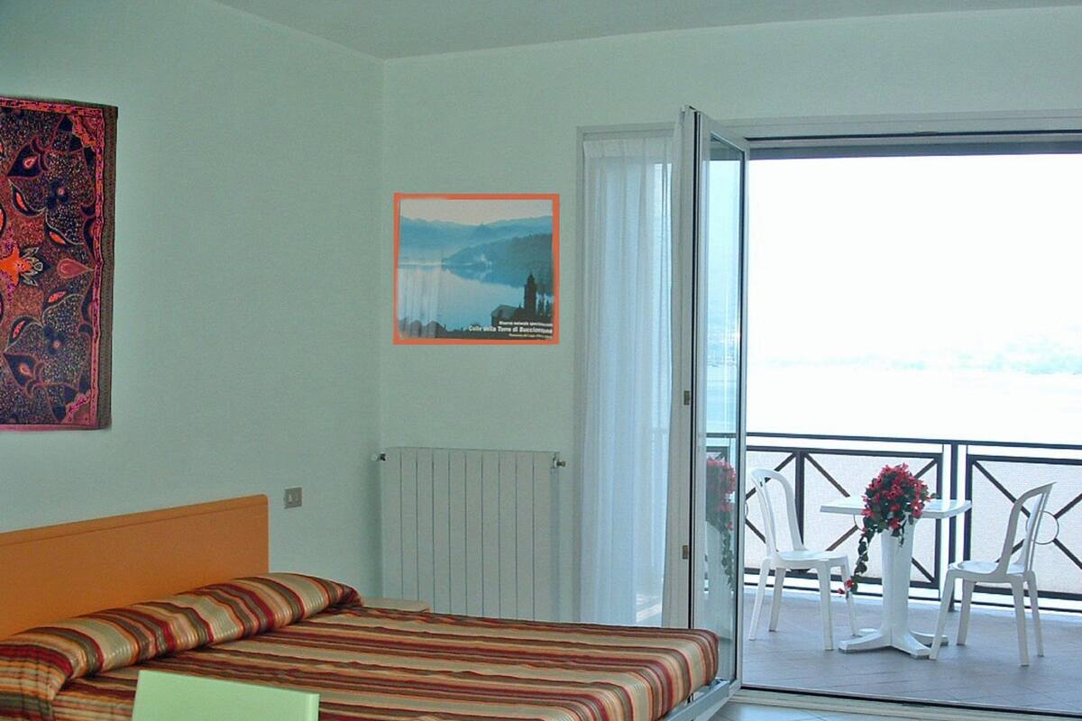 A hotel room with a bed, a chair, a table, a radiator, a picture on the wall, and a balcony with chairs and a table.