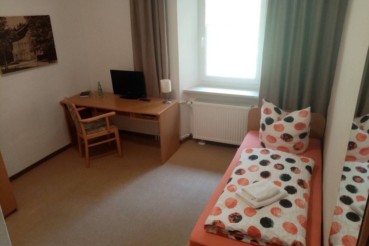 A small room with a bed, a desk, a chair, a window, and a radiator.