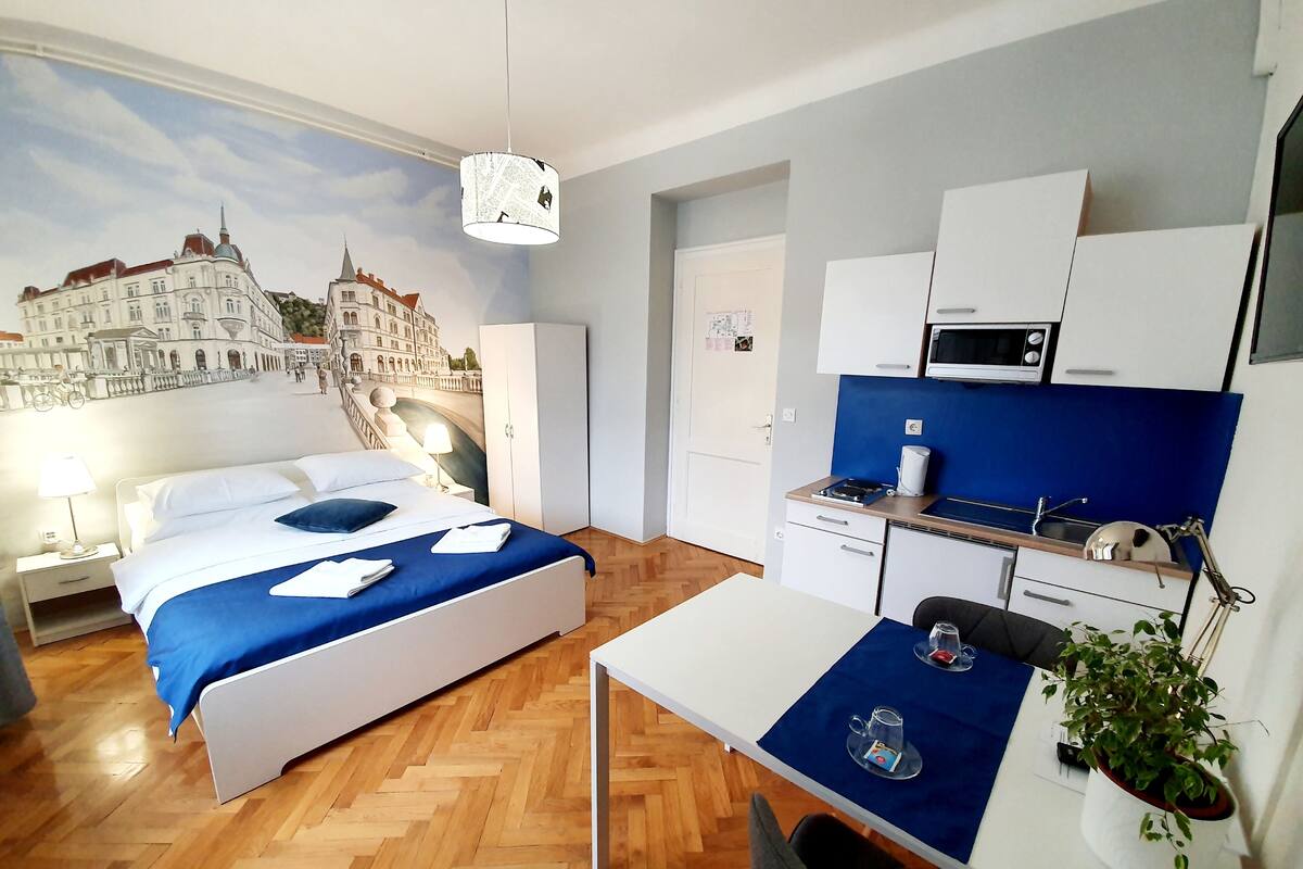 Une chambre d’hôtel équipée d’un lit, d’un bureau, d’une kitchenette et ornée d’une fresque représentant un paysage urbain.