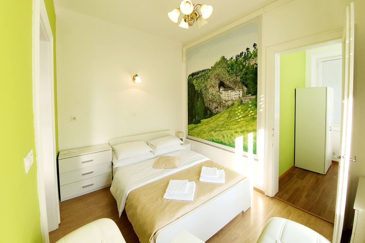 Une chambre à coucher comprenant un lit, des tables de chevet, une commode, une fenêtre donnant sur un château et une porte menant à une autre pièce.
