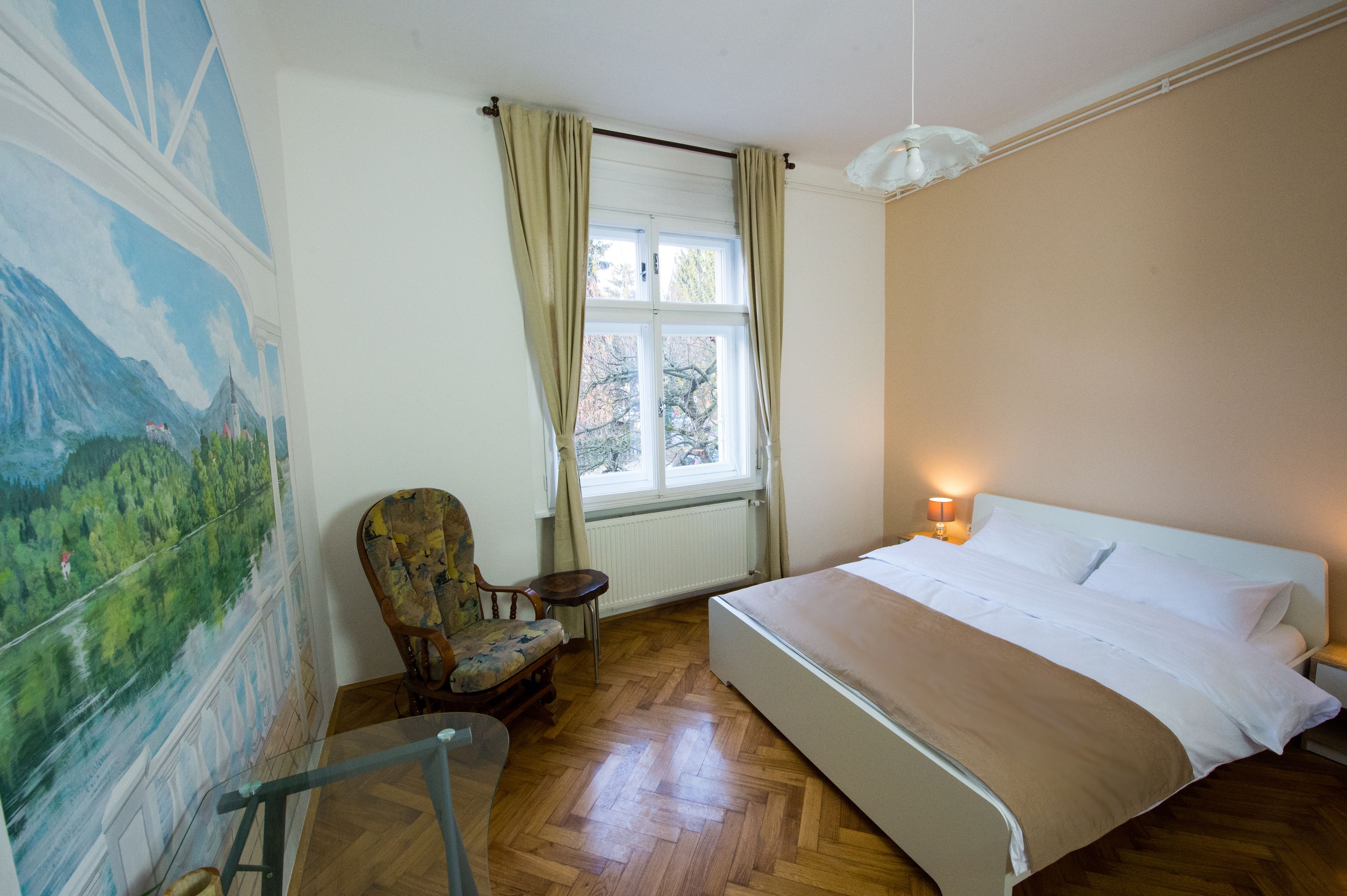 Un dormitorio con una cama, una silla, una ventana con cortinas, un radiador y un cuadro en la pared.