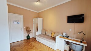 Una habitación con piso de madera en patrón de espiga, una cama con cajones, un escritorio con silla, un televisor montado en la pared y una puerta con un tablón de anuncios.