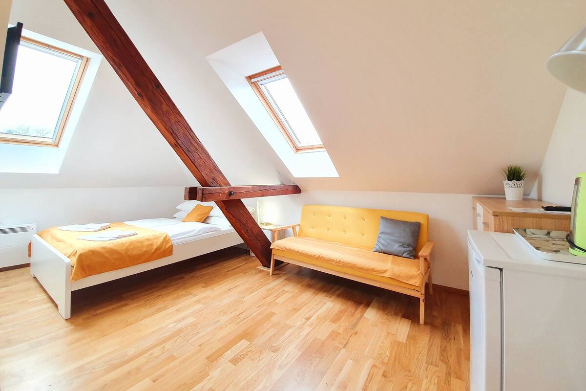 Une chambre située dans un espace de type « loft », avec un plafond en pente, des poutres en bois, deux lits, un canapé, une petite table et une plante en pot.