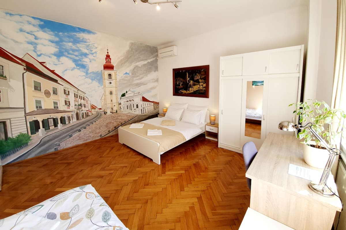 Une chambre d’hôtel avec un lit, un bureau doté d’une lampe, une plante en pot et une fresque représentant une scène de ville sur le mur.