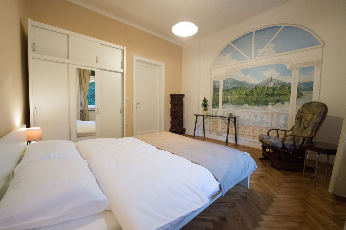 Une chambre à coucher avec un grand lit, une chaise, une petite table et une vue sur un paysage par la fenêtre.