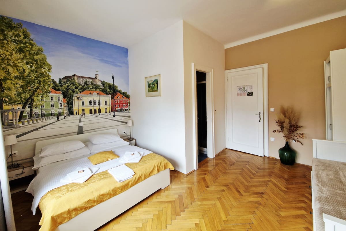 Une chambre d’hôtel avec un lit, un tableau représentant une scène de ville, un plancher en bois et une porte donnant sur une autre pièce.