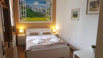 Habitación de hotel con cama, dos lámparas de noche, una mesita de noche, ventana con vistas a la montaña y una pequeña mesa con una planta.