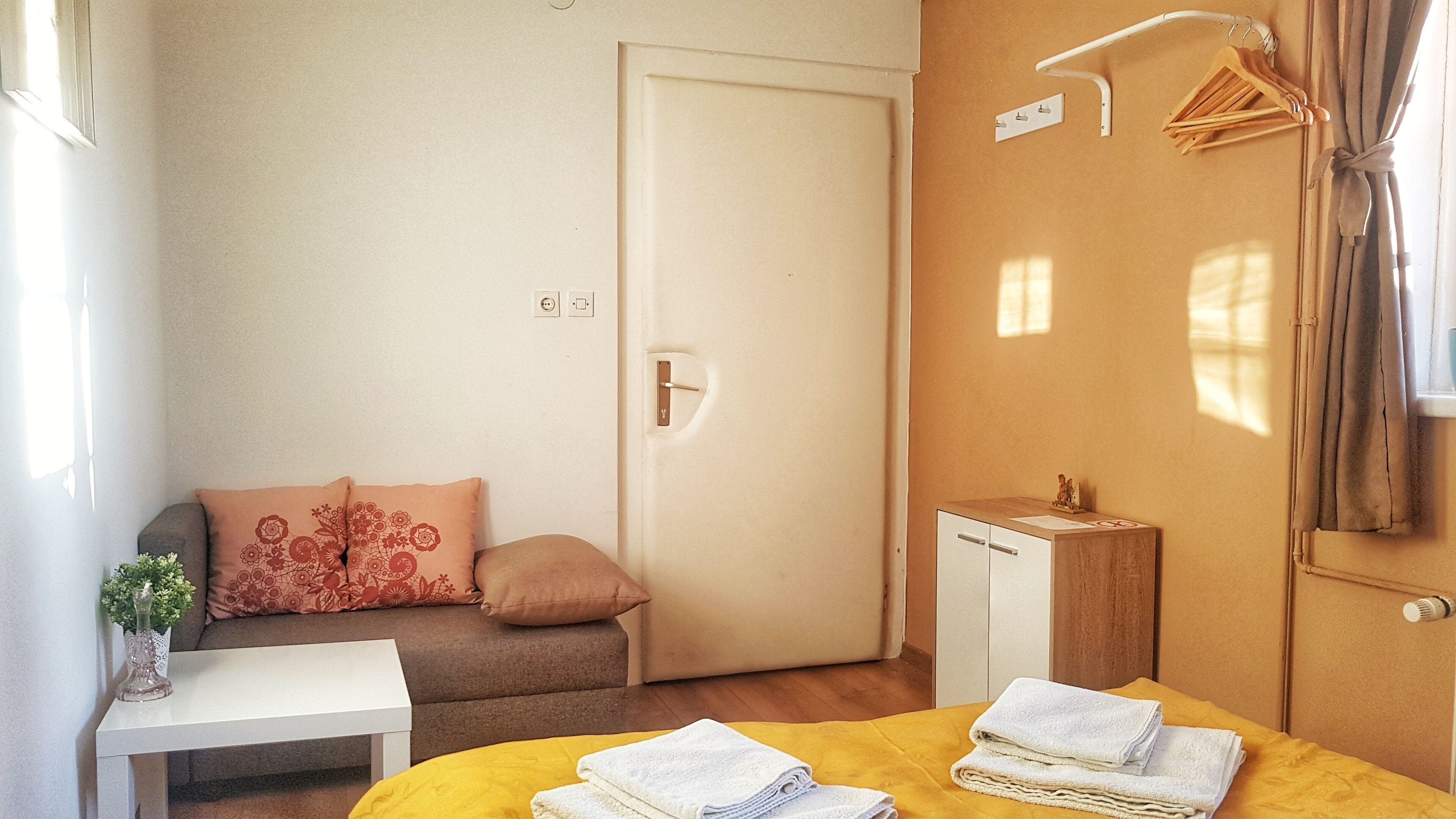 Habitació petita i ben ordenada, amb llit, taula de nit, sofà amb coixins, armari i petit moble.