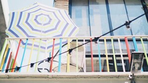 Un balcón con un paraguas a rayas, una silla y una barandilla con barras verticales de colores.