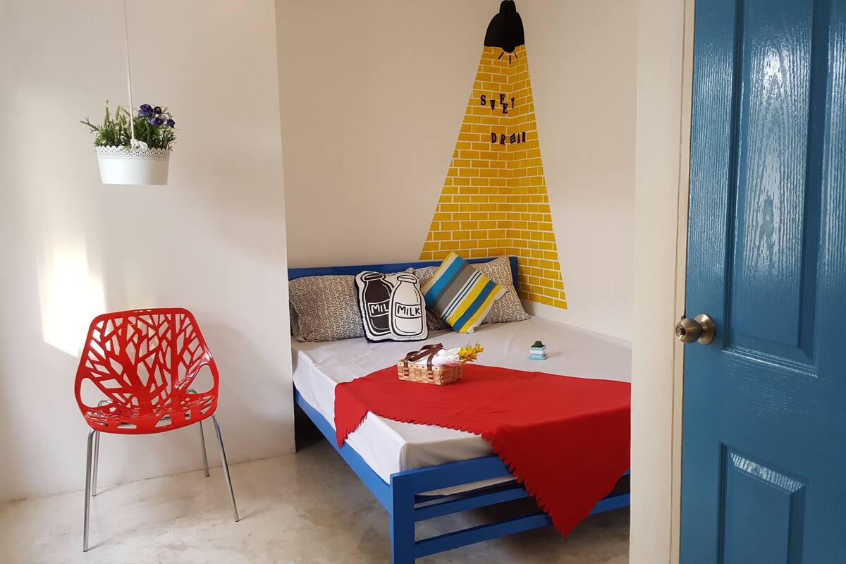 A small, neatly arranged room with a bed, a red chair, a hanging plant, and a blue door.