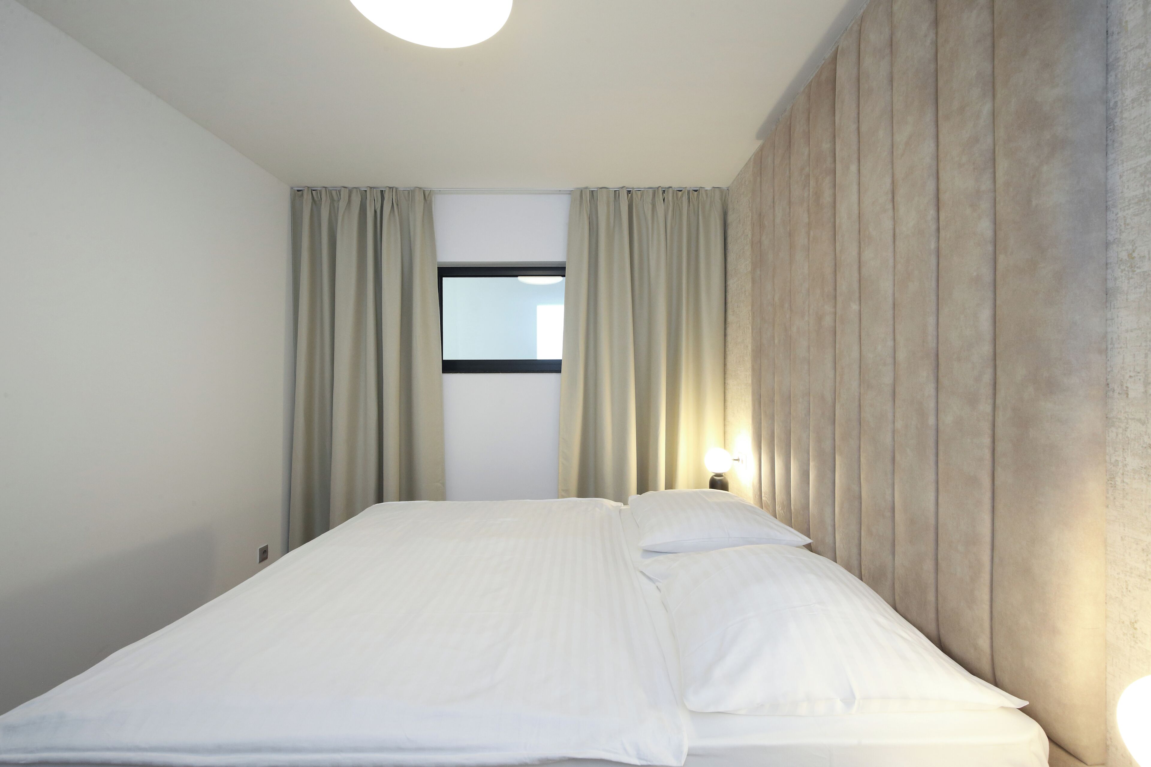 Hotellihuone, jossa on sänky, verhot, yöpöytä ja seinään kiinnitetty valaisin.