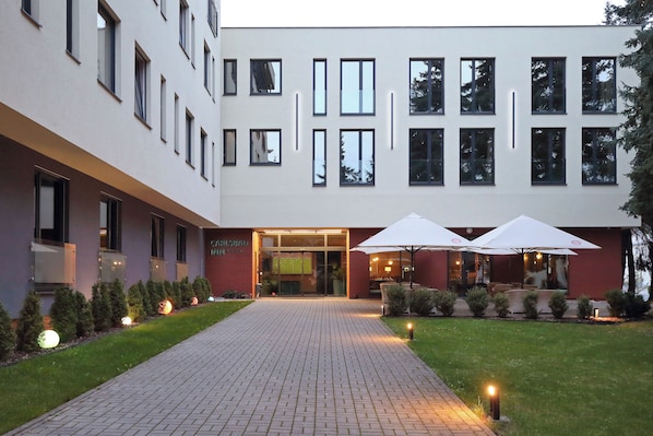 A hotel entrance with a paved walkway, outdoor seating area with umbrellas, and a well-maintained lawn.