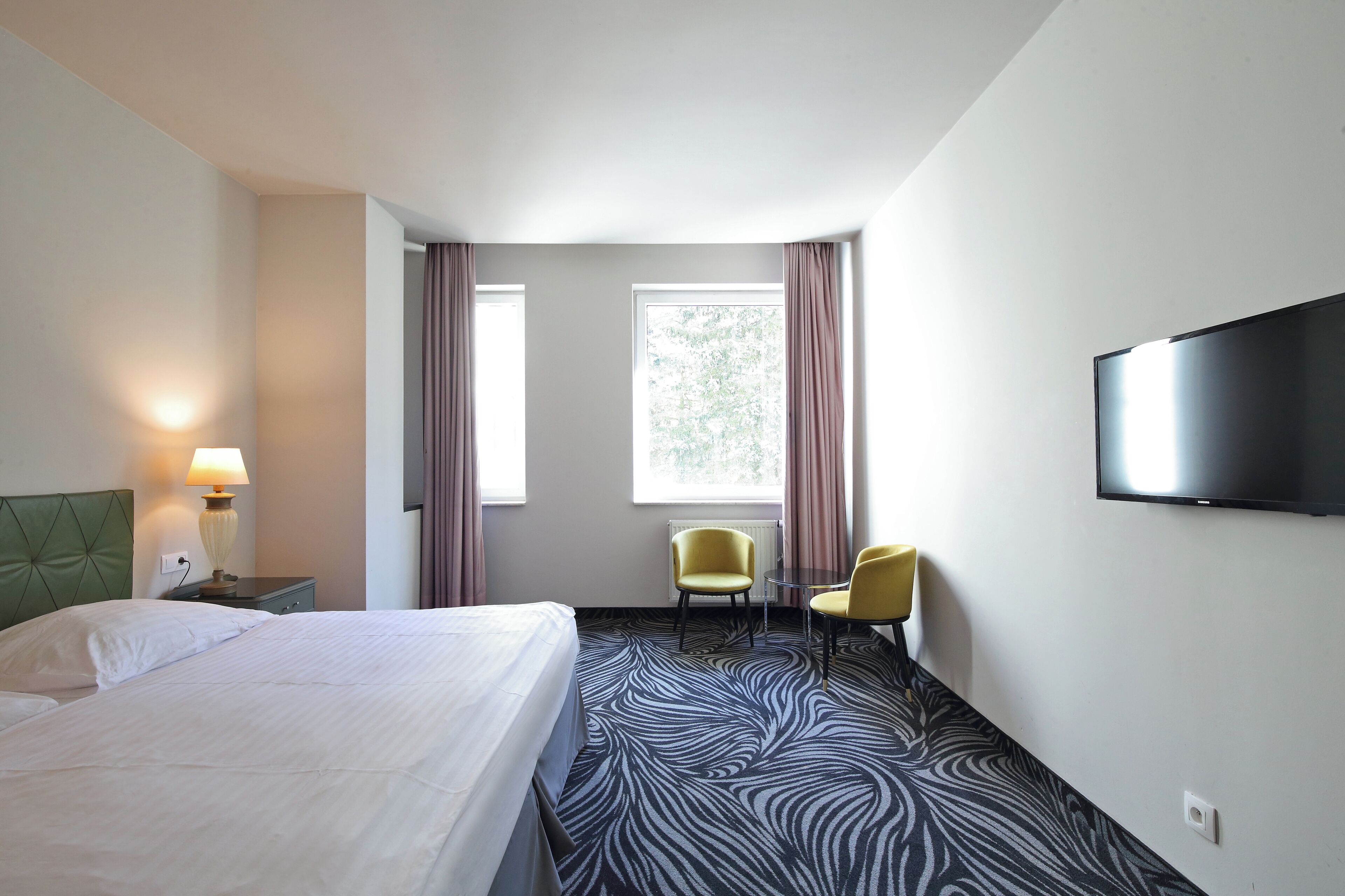 A hotel room with a bed, two chairs, a TV, and a window with curtains.