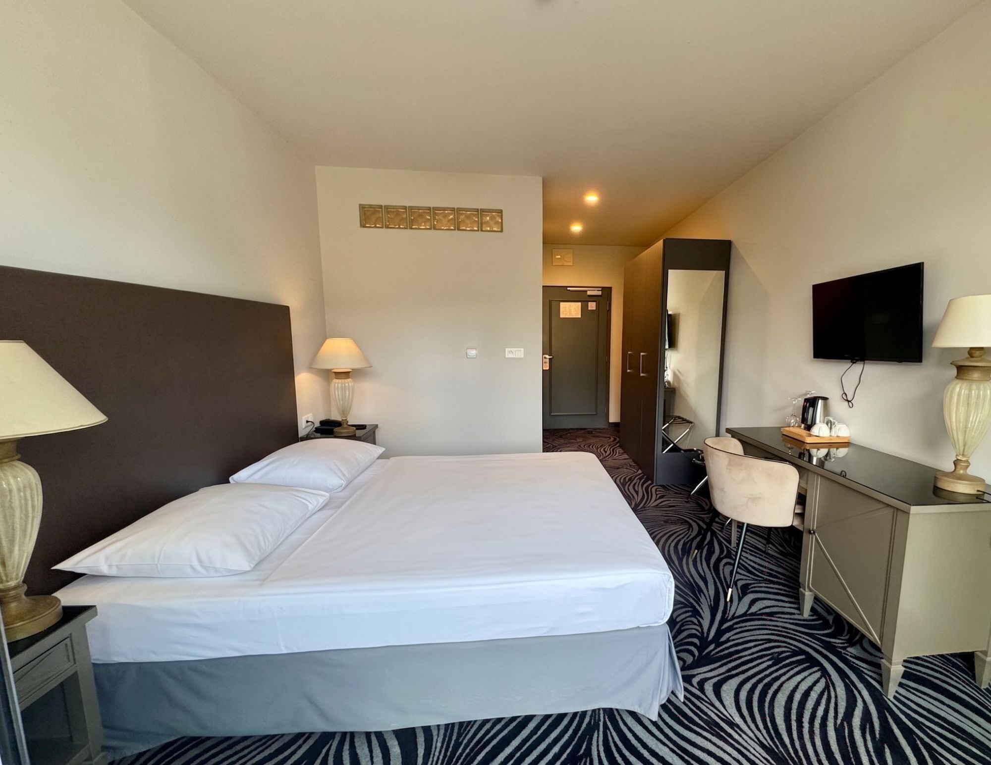 A hotel room with a large bed, bedside lamps, a desk with a mirror, a television, and a zebra-patterned carpet.