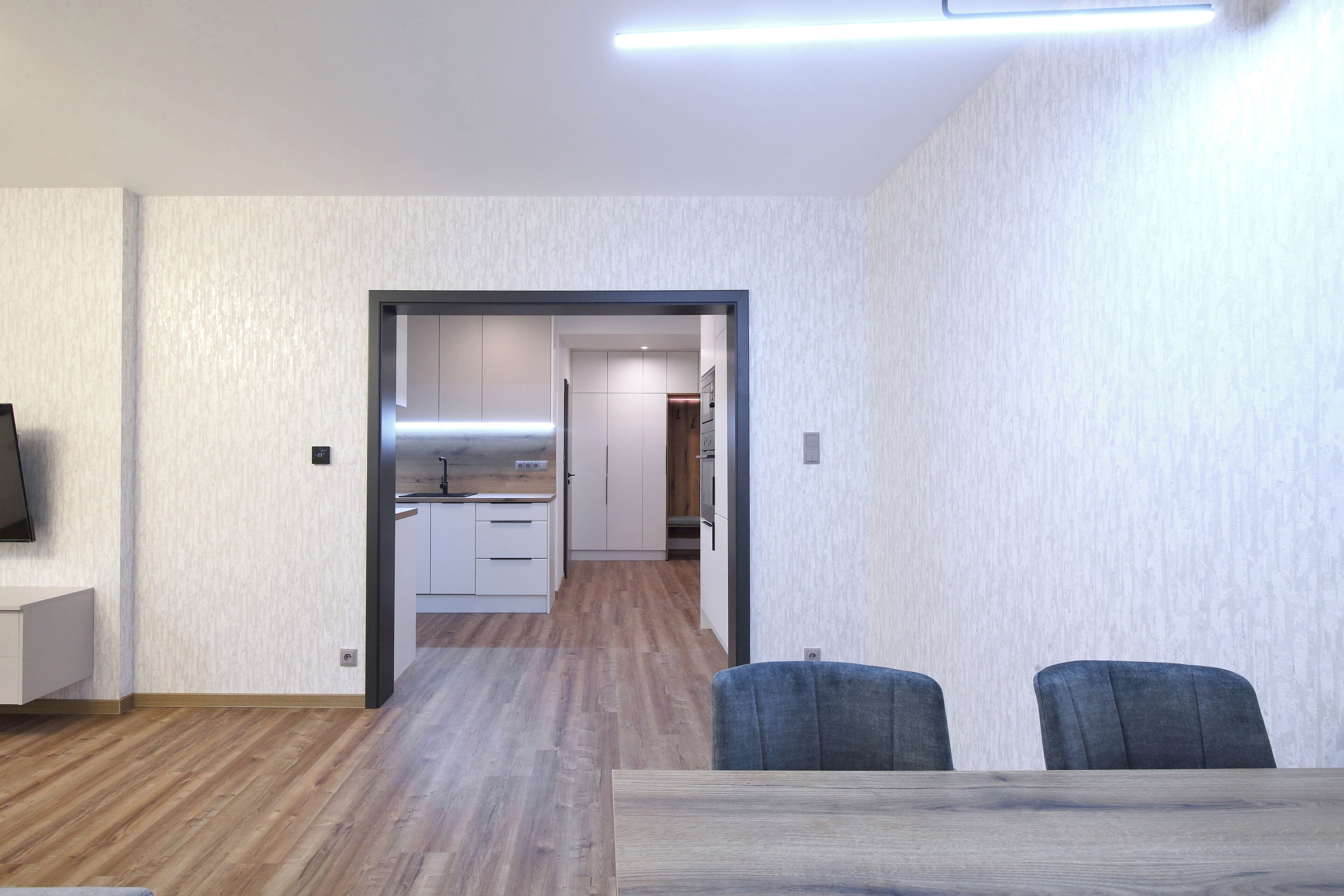 Un interior moderno con piso de madera, una zona de comedor con dos sillas y una cocina visible a través de un vano abierto.
