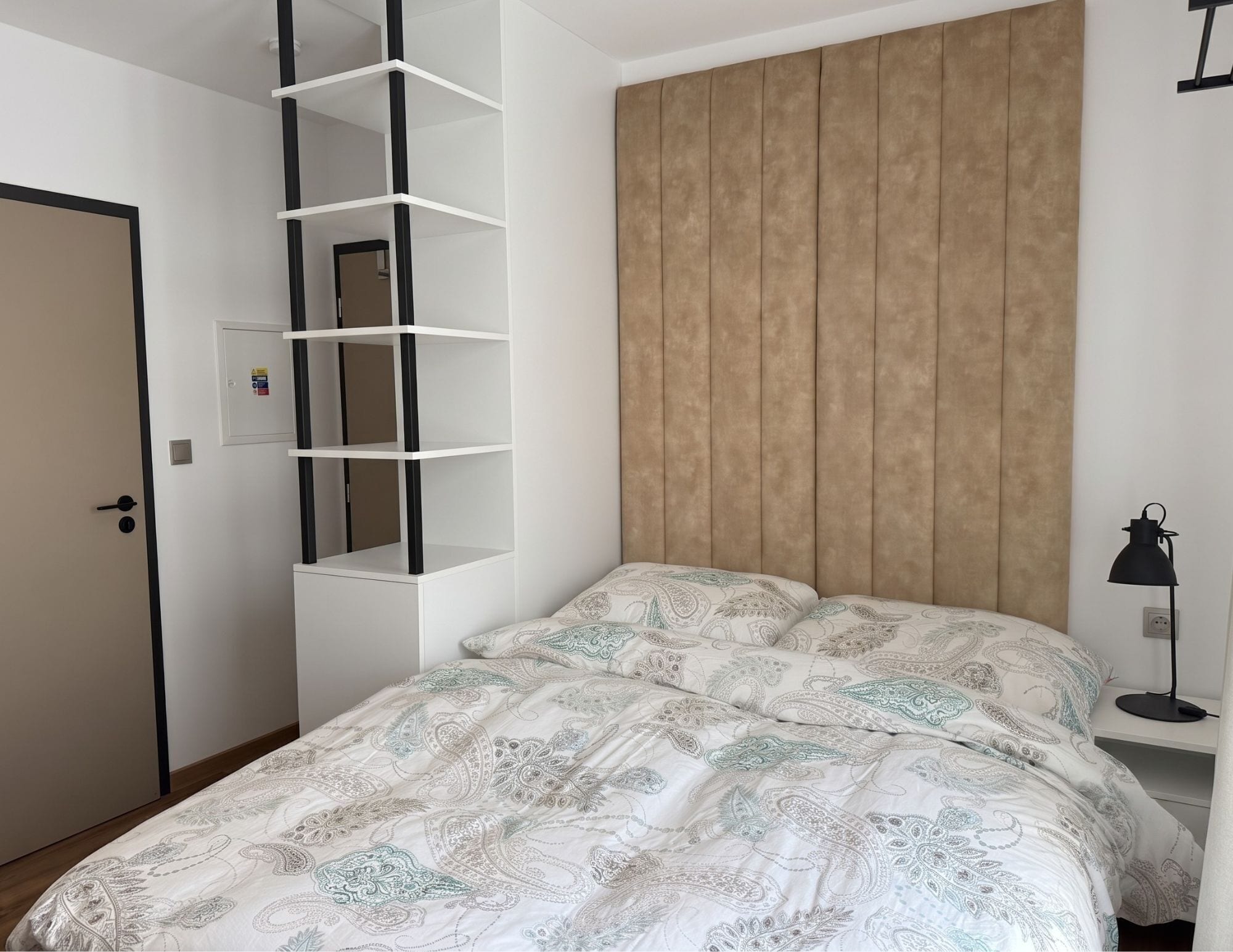 A bedroom with a bed, a wardrobe, a shelf unit, a bedside table with a lamp, and a door.
