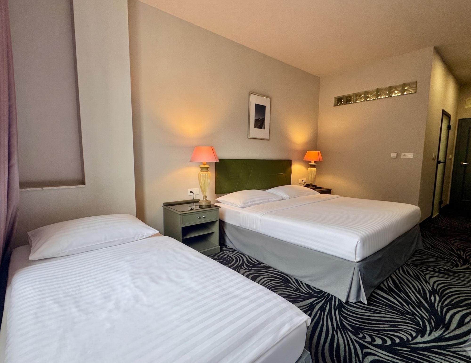 Habitación de hotel con dos camas, cabecero verde, lámparas de noche, un cuadro en la pared y alfombra con estampado de cebra.