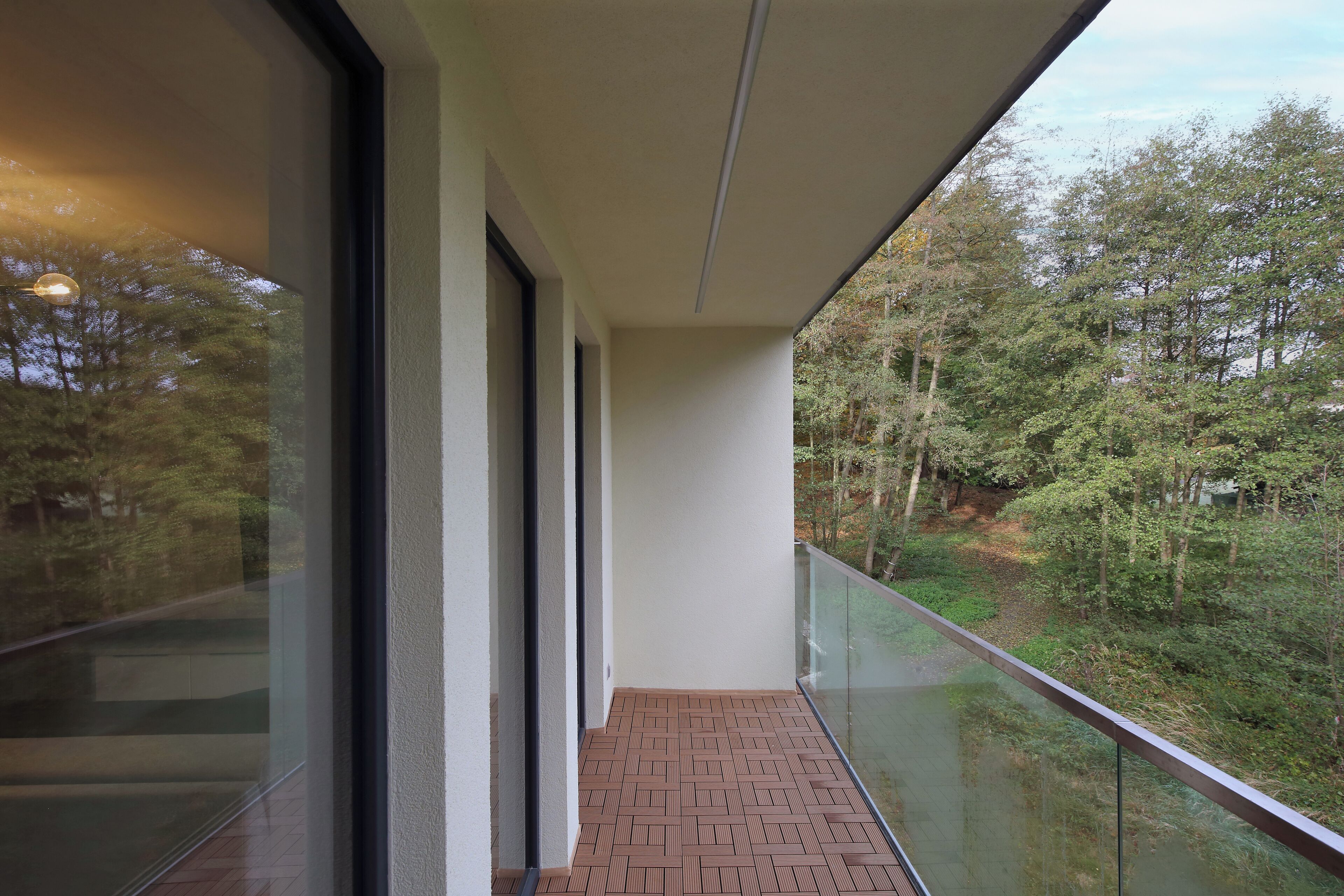 Un balcon doté d’une balustrade en verre, d’un sol en briques et offrant une vue sur une zone boisée.