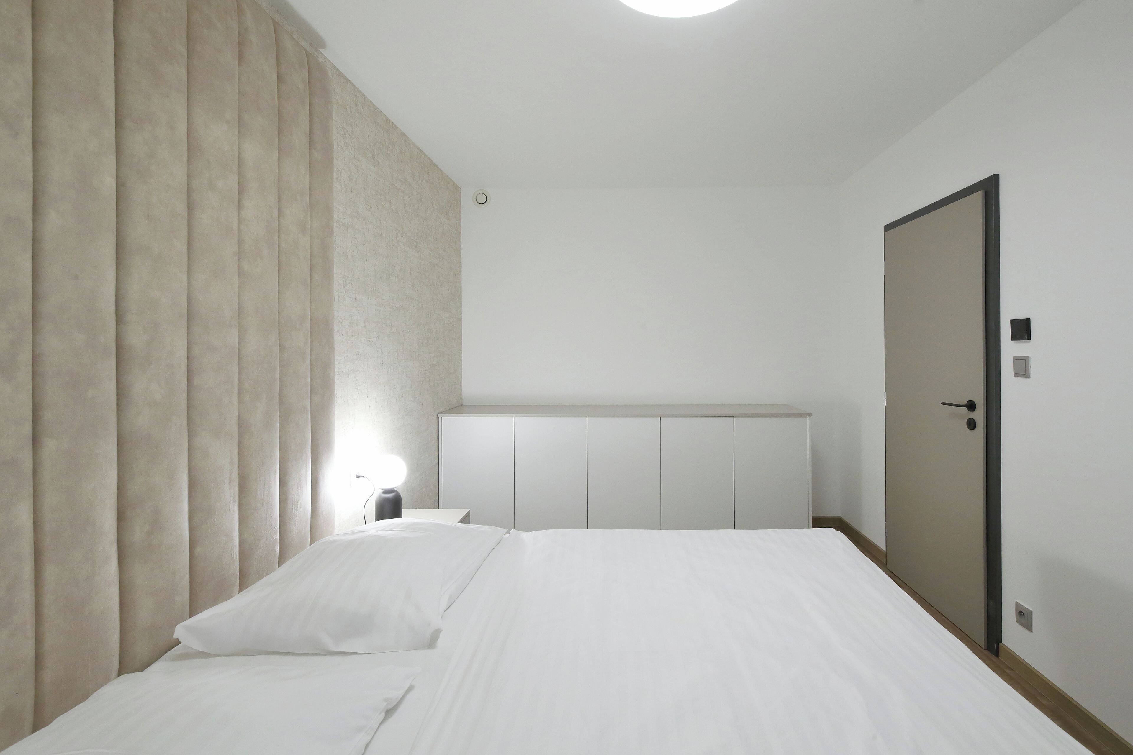 Un dormitorio con cama, mesita de noche, luz de pared y una puerta.