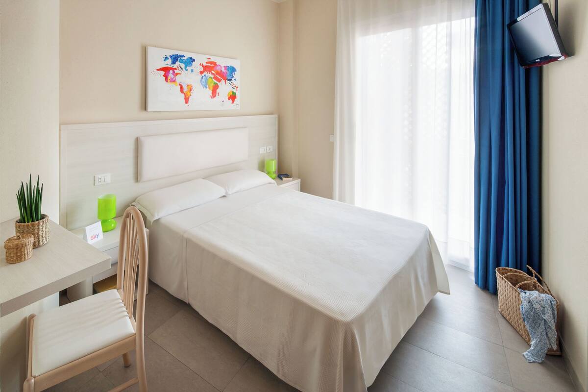 Una habitación de hotel con cama, escritorio, silla, un cuadro de un mapa mundial, televisor y cortinas.