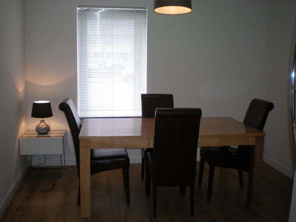 A dining area with a wooden table and four chairs, a small side table with a lamp, and a window with blinds.