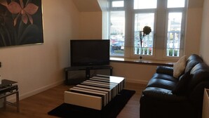 A modern living room with a black leather sofa, a flat-screen TV, a striped ottoman, and a large window with blinds.