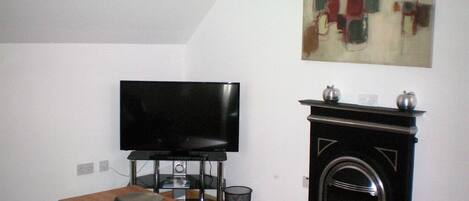 Uma sala de estar com televisão de tela plana em um suporte, mesa de centro de madeira, lareira e uma poltrona verde.
