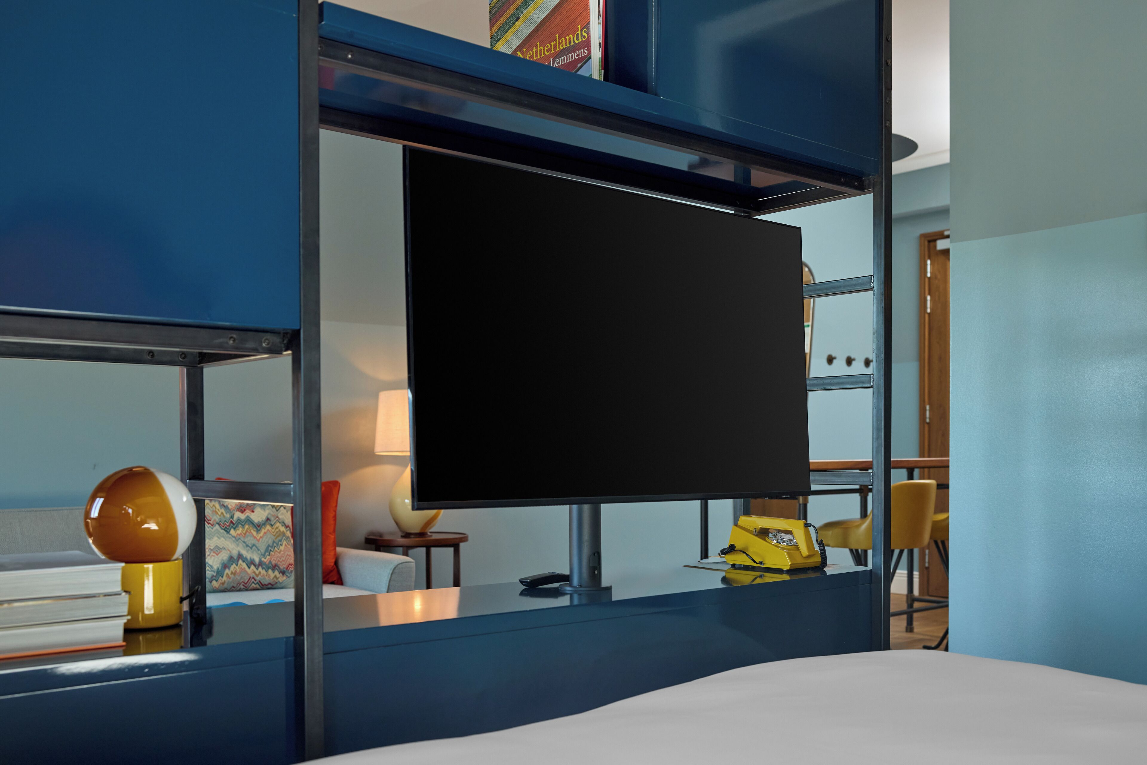 Ein modernes Hotelzimmer mit einem großen Flachbildfernseher an einer blauen Wand, einem Bett mit weißer Bettwäsche, einem kleinen Schreibtisch mit einem gelben Telefon und einem farbenfrohen Kissen auf der Couch.