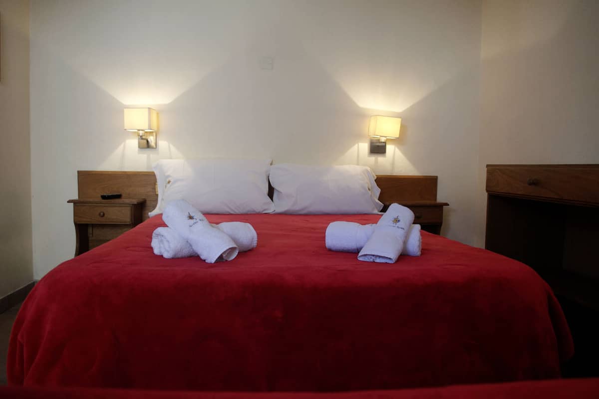 Habitación de hotel con una colcha roja, dos almohadas blancas, dos mesitas de noche con lámparas y dos toallas enrolladas sobre la cama.