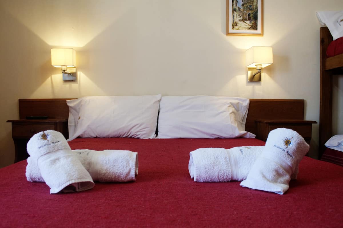 Habitación de hotel con alfombra roja, dos almohadas blancas, dos mesitas de noche con lámparas y dos toallas sobre la cama.