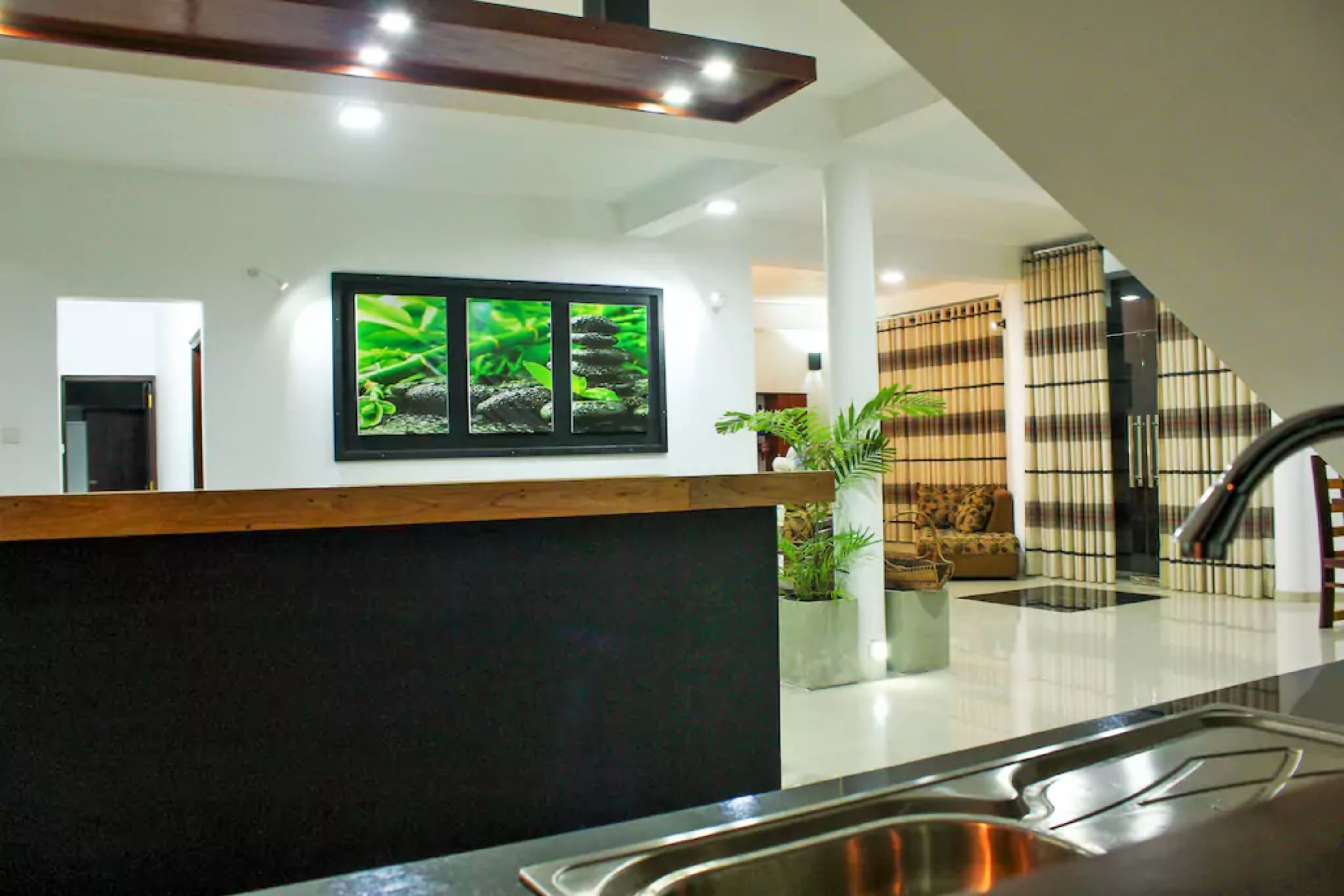 A modern kitchen with a central island, a staircase, and a living area with a sofa and plants.