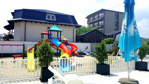 A playground with slides and a climbing structure, surrounded by a white fence, with buildings in the background.