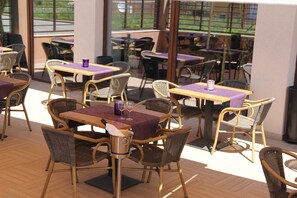 A restaurant with wicker furniture and purple tablecloths, large windows, and a view of the outdoors.
