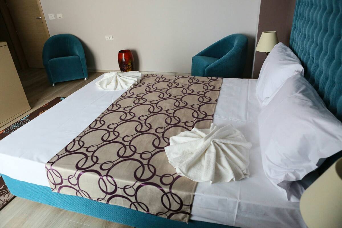 A hotel room with a bed, two blue armchairs, a vase, and a lamp.