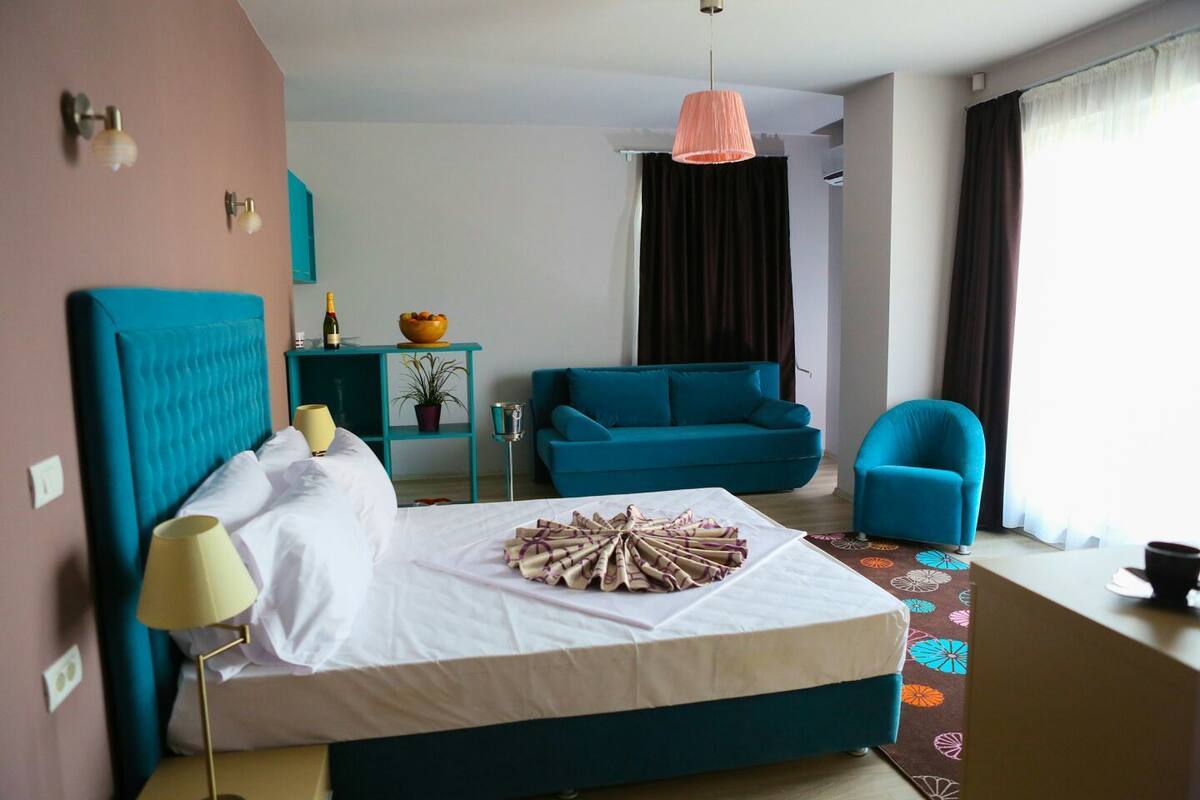 A hotel room with a bed, a teal sofa, a blue armchair, a small table with a plant, and a window with curtains.