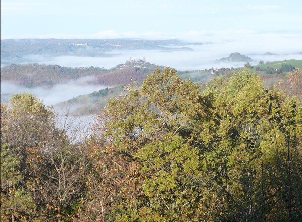A mountainous landscape with dense foliage and a distant view of a castle-like structure on a hill. - Bout De Cote (Sousceyrac-en-Quercy)