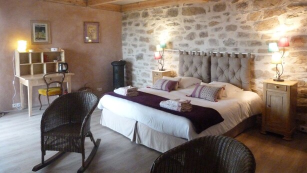 Une chambre avec un mur en pierre, un grand lit avec des oreillers, un fauteuil à bascule, un bureau avec une chaise et des lampes de chevet.