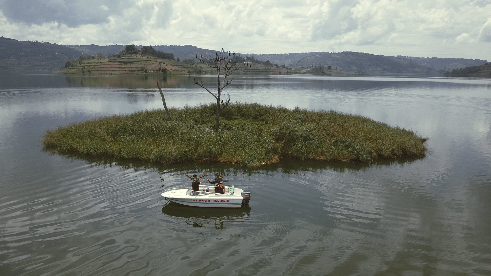 Un bote llamado "LAK BUNYONI ROCK RESORT" sobre aguas tranquilas, con una pequeña isla y colinas al fondo.