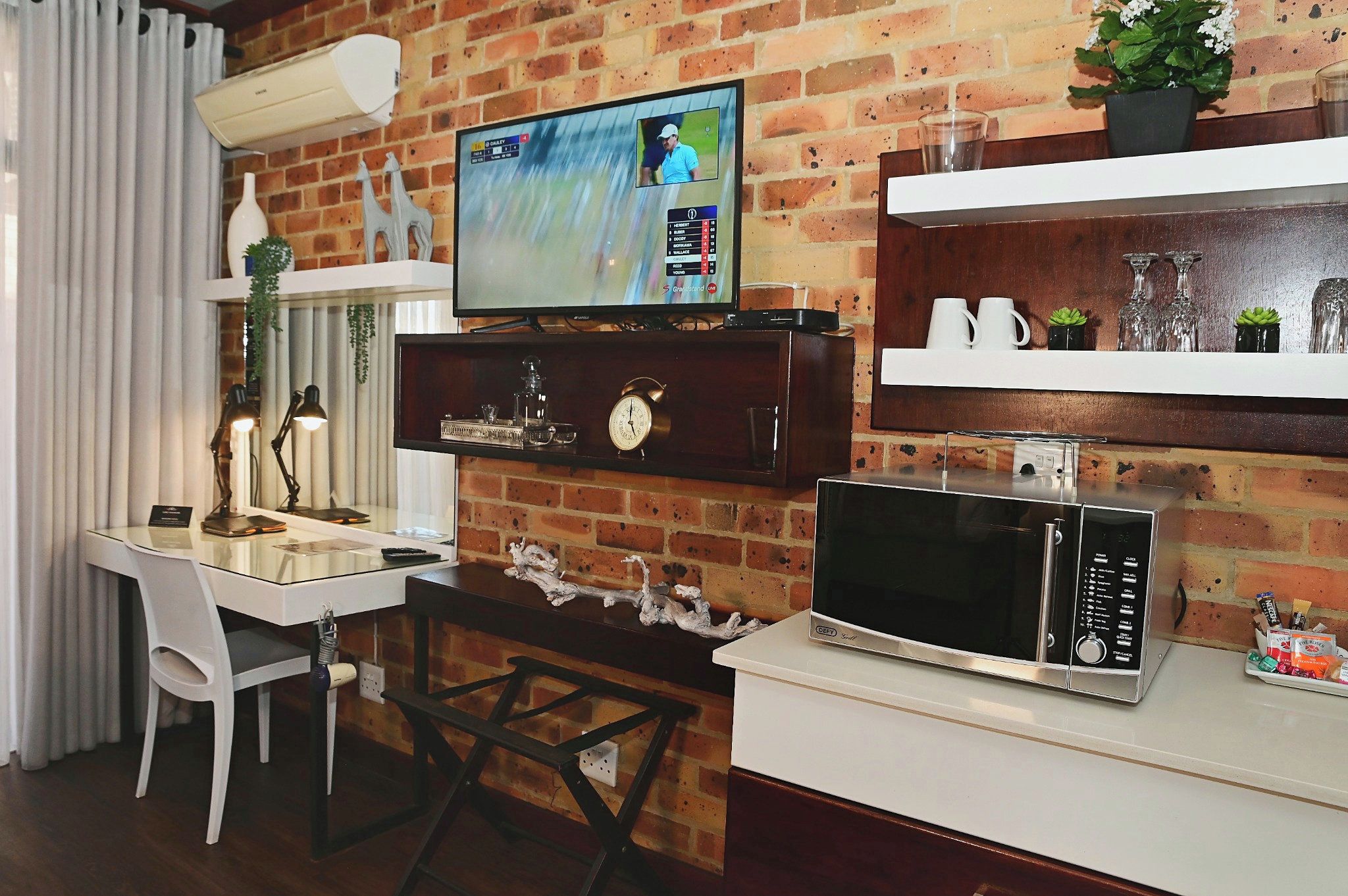 Un espacio habitable compacto con un televisor montado en una pared de ladrillo, una mesa de comedor con sillas, un microondas y estantes con objetos decorativos.