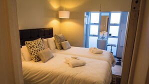 A hotel room with two beds, a headboard, a bedside table, a lamp, and a bathroom visible through an open door. - Grey Manor Guesthouse (Potchefstroom)