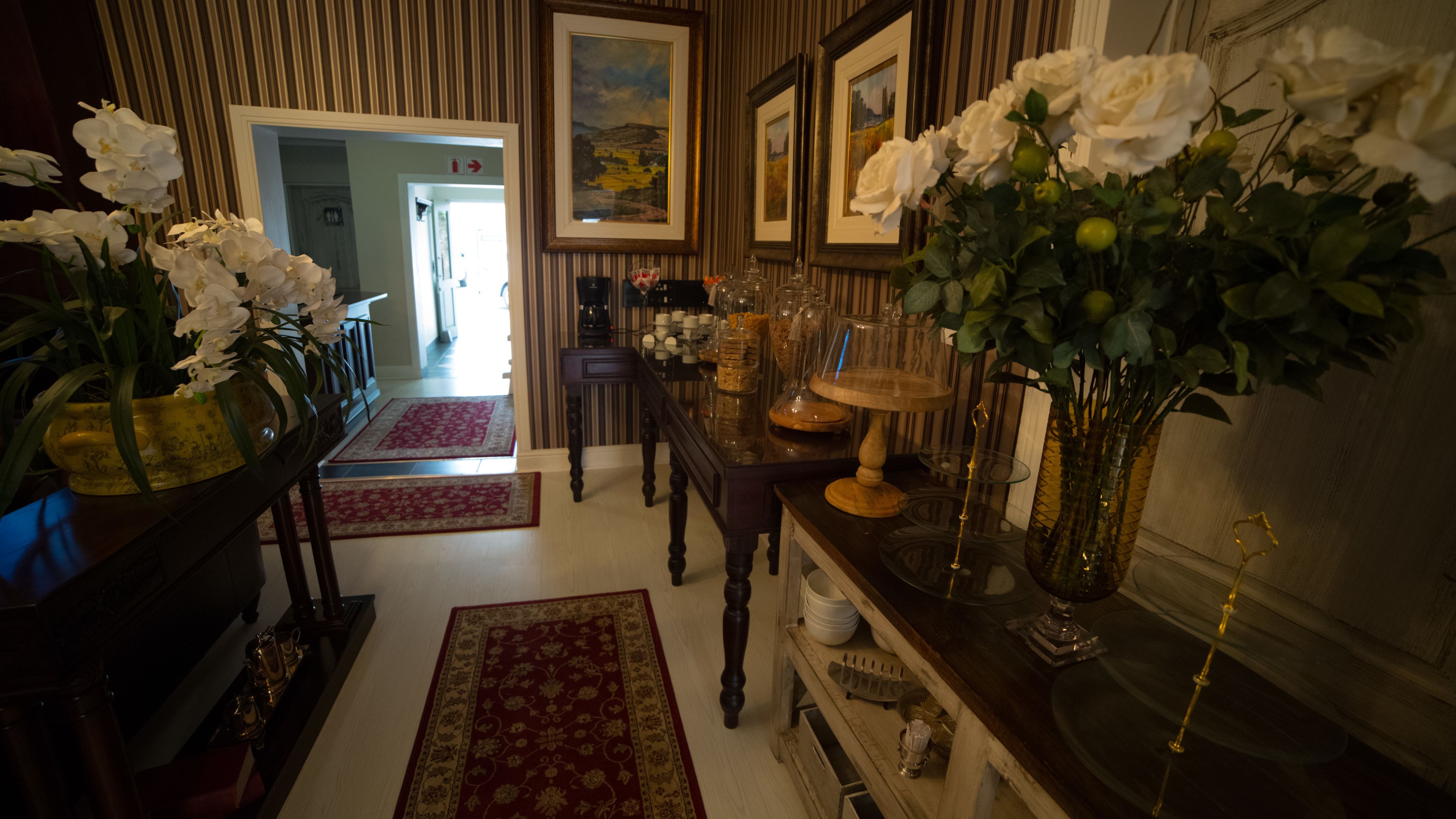 A well-lit hallway with a wooden floor, a red and gold patterned rug, a wooden table with glassware and a vase of white flowers, and framed pictures on the wall.