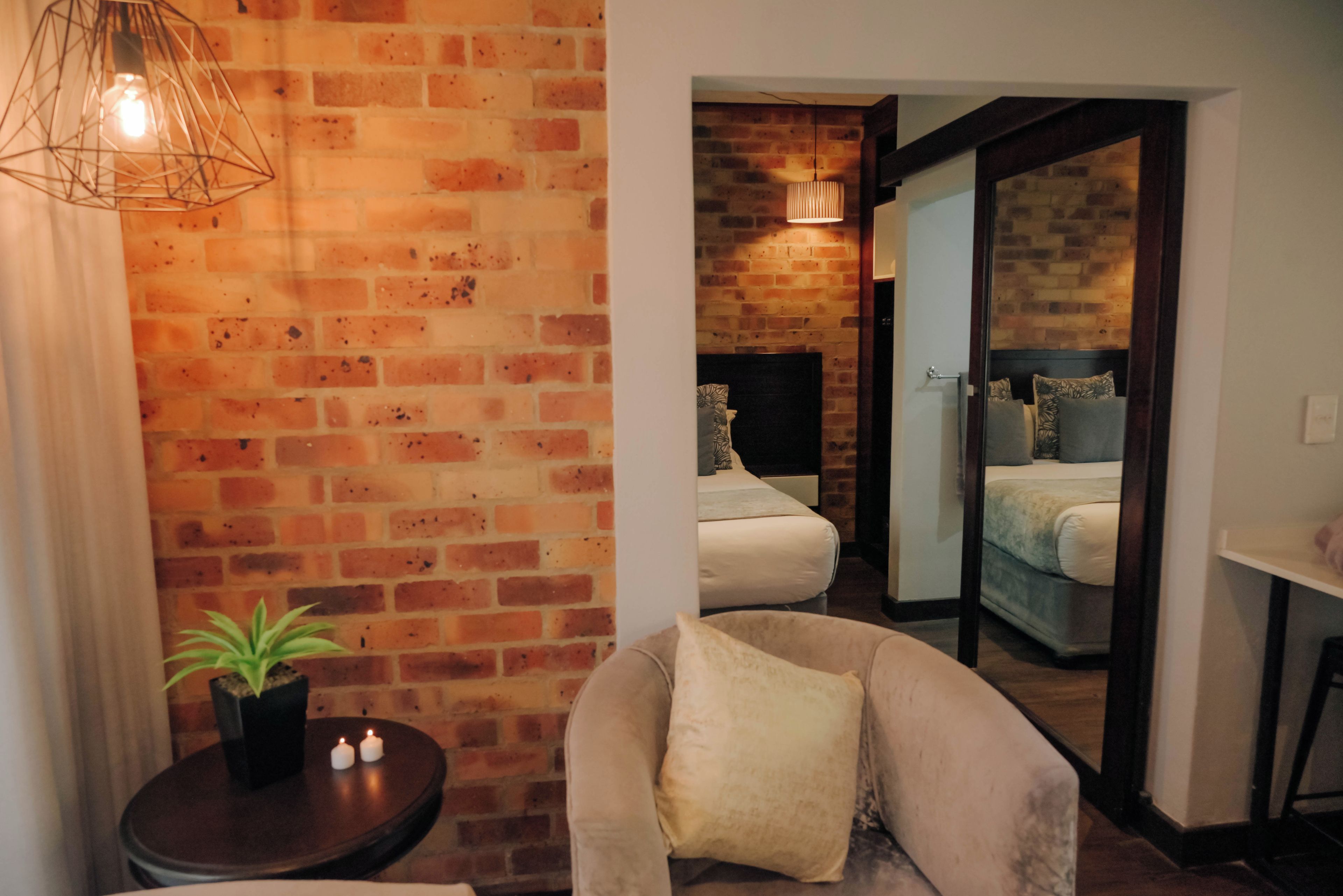 A room with a brick wall, a bed with a headboard, a chair with a cushion, a small table with a plant, and a hanging light fixture.