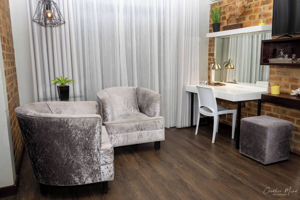 Ein modernes Zimmer mit einem grauen Sessel, einem weißen Schreibtisch und einem kleinen Hocker. Eine Kühlpflanze und eine Pendelleuchte sind ebenfalls vorhanden.