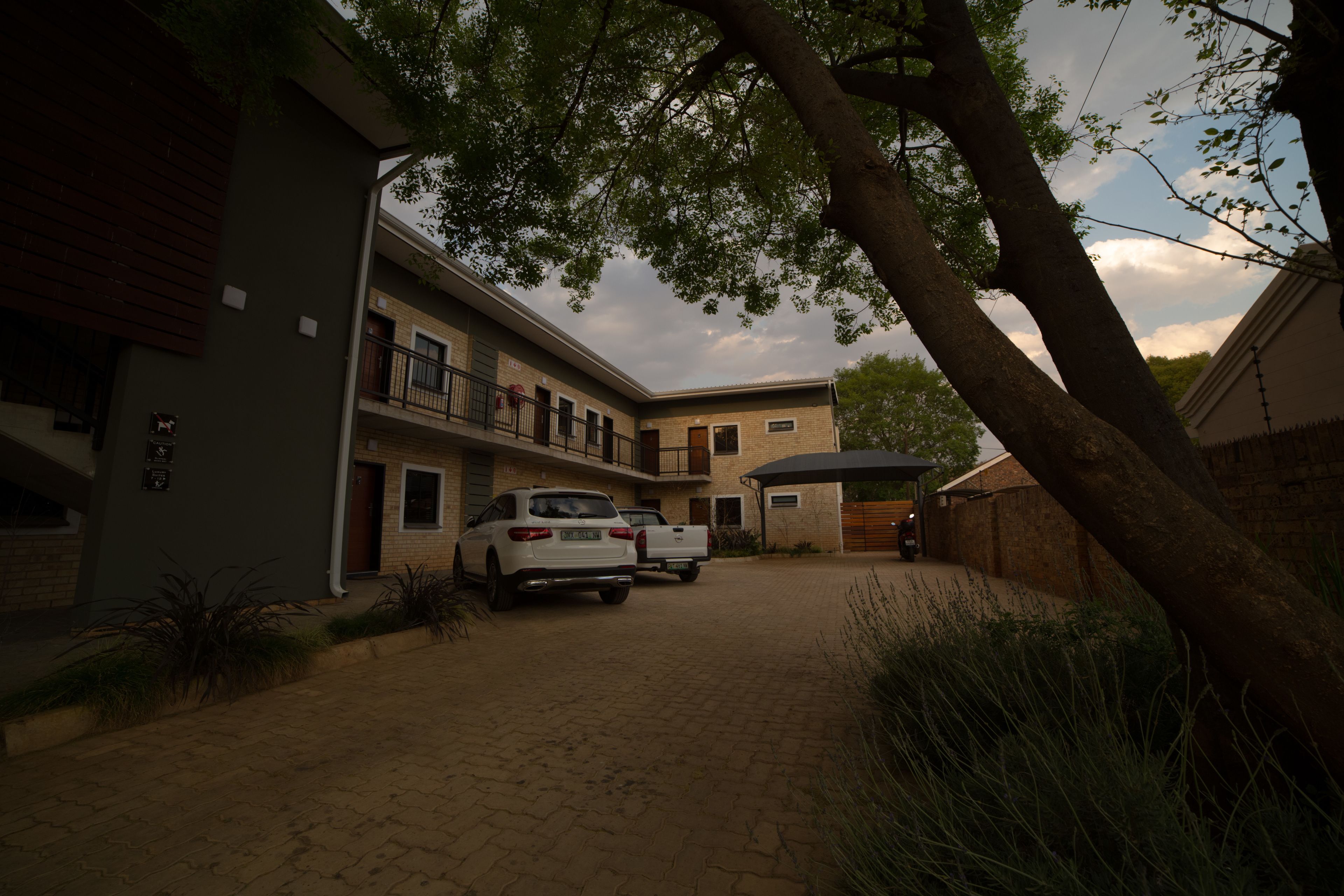 Um edifício residencial com entrada pavimentada, carros estacionados e uma árvore ao primeiro plano.