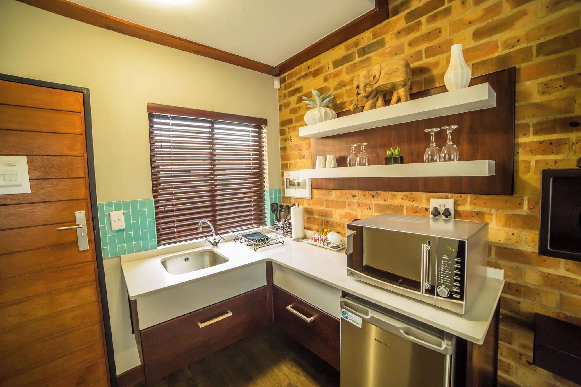 A compact kitchen with a sink, a microwave, and a dishwasher. There are wooden cabinets, a window with blinds, and a brick wall.