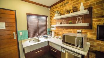 A compact kitchen with a sink, a microwave, and a dishwasher. There are wooden cabinets, a window with blinds, and a brick wall.