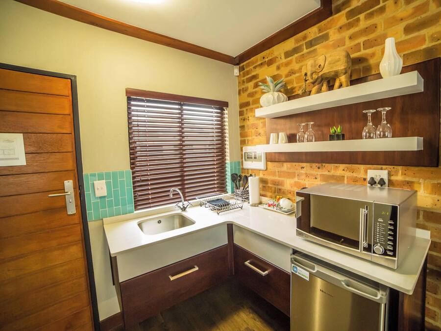 A compact kitchen with a sink, a microwave, and a dishwasher. There are wooden cabinets, a window with blinds, and a brick wall.